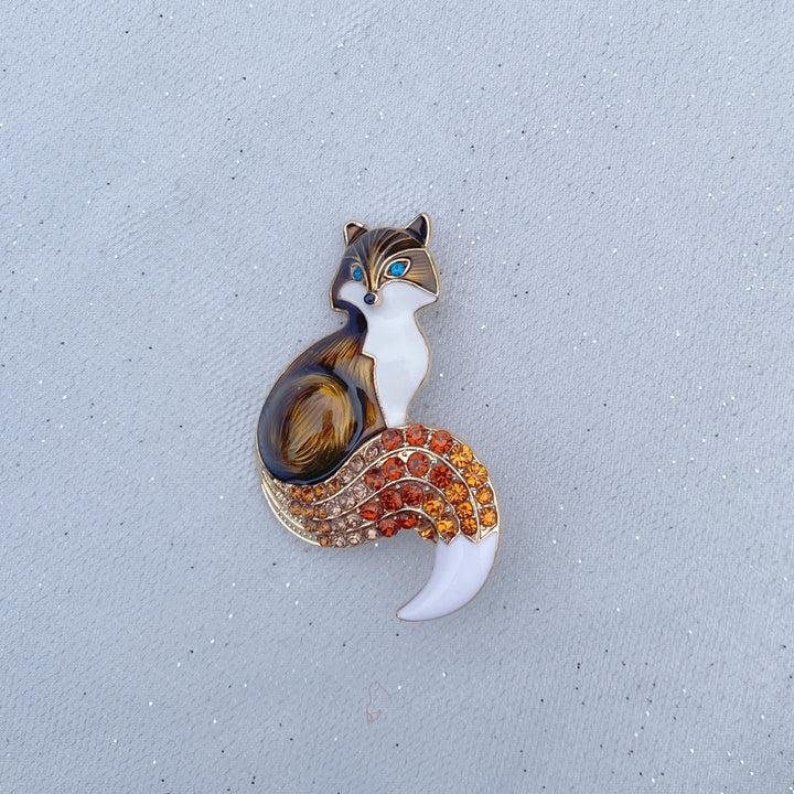 Brown and white fox brooch with blue eyes and an orange crystal tail.